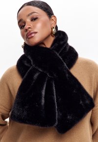 Kvinde med slicked-back hår iført guld hoops, et sort faux fur tørklæde og en tan sweater foran en hvid baggrund.