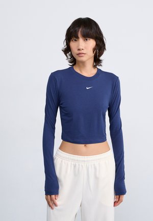 Marineblaues, hochgeschlossenes Langarmshirt mit Rundhalsausschnitt und weißem Nike-Logo auf der Brust, kombiniert mit weißen Hose mit elastischem Bund.