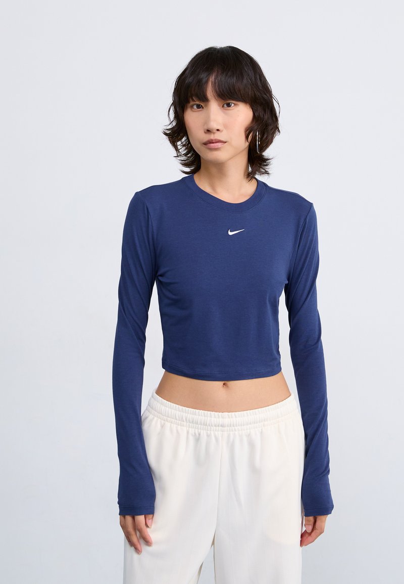 Tamsiai mėlyna, trumpa ilgio, ilgomis rankovėmis marškinėliai su apvalia iškirpte ir balta Nike logo ant krūtinės, derinami su baltais elastingais juosmens kelnėmis.