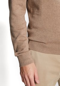 Homme portant un pull beige à manches longues et un pantalon beige, la main détendue reposant sur le côté.