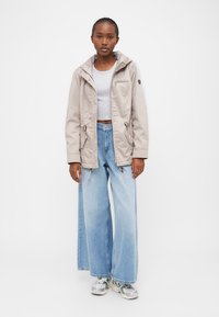 Veste beige à capuche avec taille à cordon, poches avant et fermeture éclair. Portée sur un top court gris et un jean baggy bleu clair.