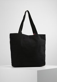 Bolsa de lona negra con dos asas superiores, forma estructurada, textura suave y un pequeño acento colorido en un lado.