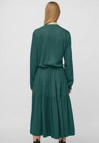 Pitkähihaisessa, tummanvihreässä mekossa on sileä kangas. Mekossa on vyötärönauha ja kerroksellinen hameosa. Takakuvassa helma ulottuu juuri kalleilta alapuolelle.