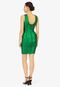 Robe verte ajustée avec un dos décolleté, motif texturé et fermeture éclair. Chaussures à talons noires avec des accents arrondis complètent le look.