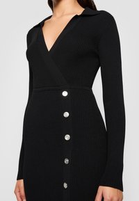 Femme portant une robe en maille côtelée noire, à manches longues, col en V profond, col et une rangée de cinq boutons argentés sur le côté avant.