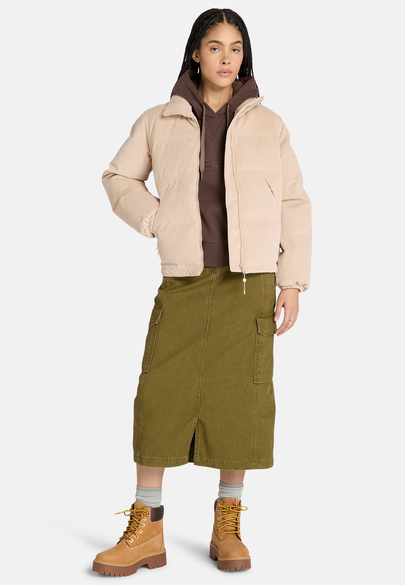 Giubbotto imbottito in corduroy beige, felpa marrone, gonna midi verde oliva e stivali color beige con accenti marroni. Presenta una chiusura lampo frontale e tasche.