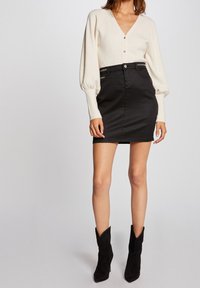 Cardigan blanco de cuello en V con mangas abullonadas y tres botones dorados, combinado con una falda mini de mezclilla negra y botas de tobillo de ante negro.