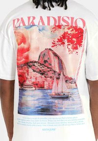 Witte t-shirt met een kleurrijke grafische afdruk van een kustlandschap met een berg, zeilboot en de tekst "PARADISIO" in het roze.