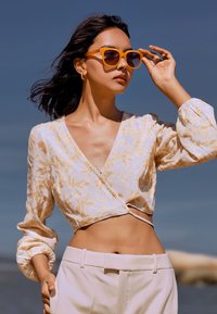 Blusa crema con patrón floral dorado, escote en V profundo y mangas abullonadas. Combinada con pantalones de talle alto en beige claro y gafas de sol naranjas.