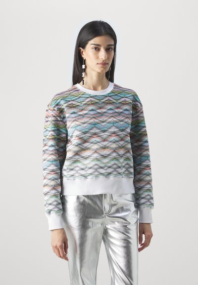Χρωματιστό πουλόβερ με μοτίβο κυμάτων σε διάφορες αποχρώσεις, με λευκό ριχτό γιακά και μανσέτες. Συνδυάζεται με γυαλιστερό ασημένιο παντελόνι.