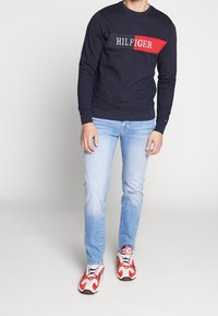 Námořnická mikina s červeným a bílým logem, modré džíny a červené a bílé tenisky. Mikina má kulatý výstřih a žebrované manžety.