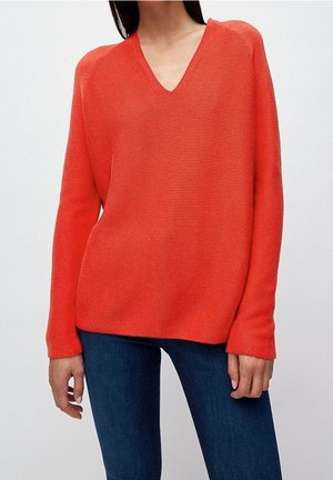 Femme portant un pull tricoté col en V à manches longues de couleur orange vif et un jean bleu foncé sur un fond clair uni.