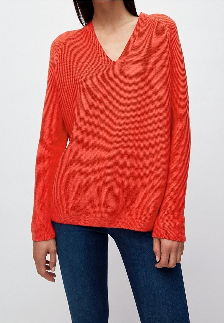 Femme portant un pull tricoté col en V à manches longues de couleur orange vif et un jean bleu foncé sur un fond clair uni.