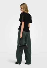 Femme souriante, portant un haut à manches courtes en dentelle noire, un pantalon large vert foncé, tenant un sac à main noir, debout devant un fond clair uni.