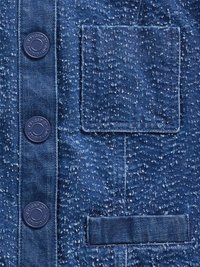 Teren de denim de culoare albastră închisă, cu un model texturat. Patru nasturi închiși la culoare pe laterale și un buzunar dreptunghiular vizibil.