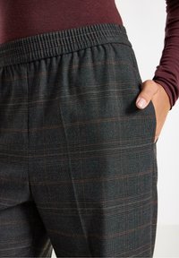 Nærbillede af en person iført mørkegrå ternede elastiske bukser med en hånd afslappet placeret i sidelommen, burgunder overdel synlig.