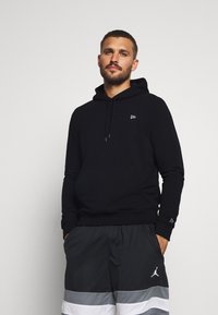 Felpa nera in cotone con tasca frontale e un piccolo logo, abbinata a pantaloni corti grigi con strisce bianche. Design semplice, vestibilità rilassata.