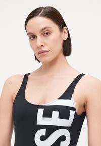 Maillot de bain noir avec de grandes lettres blanches sur le devant, bretelles fines et une texture subtile. Présente un détail blanc contrastant sur le côté.