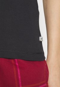Svart ribbet tank topp med en liten merkeetikett i kanten, kombinert med røde rutete bukser med gule og rosa linjer.