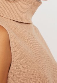 Strik turtleneck sweater i lys beige med ribbet tekstur, der har en ærmeløs design og diagonal mønster langs stoffet.