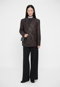 Sacou din piele maro cu rever ascuțit, un singur nasture și buzunare frontale, purtat cu pantaloni negri cu picioare largi și o geantă mică neagră.