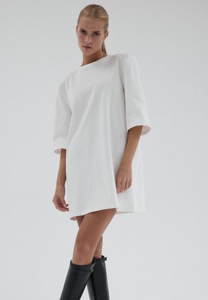 Biała bawełniana sukienka z okrągłym dekoltem, rękawami do łokcia i luźnym krojem. Noszona z czarnymi kozakami do kolan.