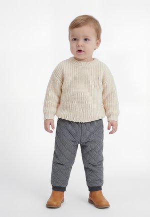 Tout-petit debout portant un pull en maille crème, un pantalon matelassé gris et des bottines marron sur fond blanc.