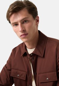 Braune Knopfjacke mit zwei Brusttaschen, Druckknopfverschlüssen und einer glatten Stoffoberfläche, kombiniert mit einem cremefarbenen Hemd.