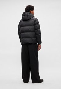 Veste matelassée noire avec capuche, présentant un design matelassé et des poignets élastiques, associée à un pantalon noir ample et des chaussures.