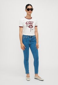 Bela bombaš t-shirt s rdečim detajli in grafiko "Cherry Coke", v kombinaciji z modrimi ozkimi jeans hlačami z navpičnimi šivi in svetlimi čevlji.