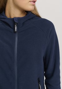 Giacca in pile blu marina con zip frontale, cappuccio e tasche laterali, caratterizzata da una superficie testurizzata e dettagli con zip a contrasto.