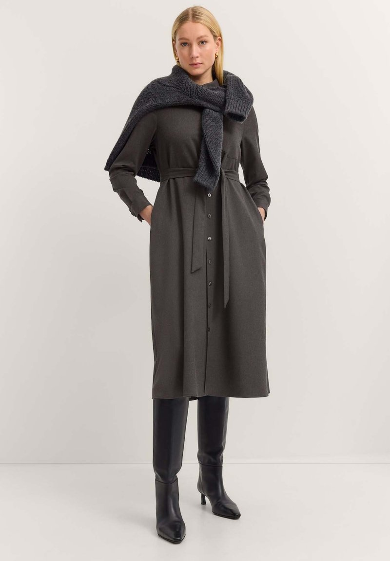 Dunkelgraues Hemdkleid mit einem Bindegürtel, langen Ärmeln und einem dunkelgrauen gestrickten Schal, der über die Schultern fällt. Schwarze knielange Stiefel.