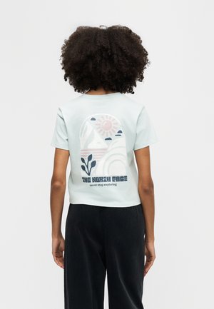 Enfant aux cheveux bouclés portant un t-shirt bleu clair avec un motif montagne, soleil et plante, ainsi que le texte « The North Face never stop exploring » au dos.