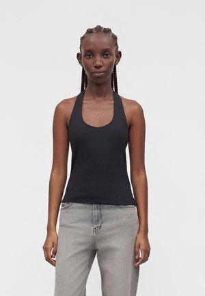 Hoikka henkilö, jolla on letitetyt hiukset, pukeutunut hihattomaan mustaan toppiin ja vaaleanharmaisiin farkkuihin, seisoo yksinkertaisella vaalealla taustalla.