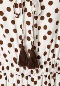 Weißes Kleid aus Stoff mit großen braunen Polka Dots, das eine geraffte Taille, Zugbänder und dekorative braune Quasten aufweist.