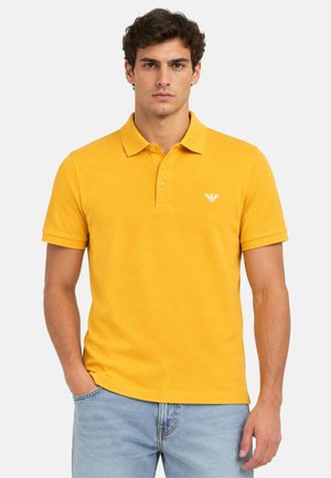 Uomo che indossa una polo gialla a maniche corte con un piccolo logo bianco sul petto e jeans azzurri chiari.