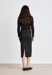Brauner Pullover kombiniert mit einem schwarzen Midirock mit Nadelstreifen, der einen Mittelschlitz am Rücken hat, und schwarzen flachen Schuhen, von hinten betrachtet.