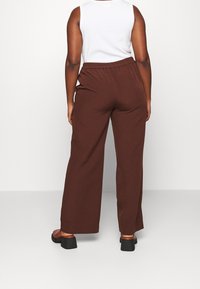 Brązowe spodnie o szerokich nogawkach widziane od tyłu, z elastycznym paskiem w talii i gładkim materiałem. Na dole widoczne sandały na obcasie.