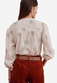 Light beige shirt with green floral embroidery, textured fabric, puffed sleeves. Paired with rust-colored corduroy pants and a tan belt.