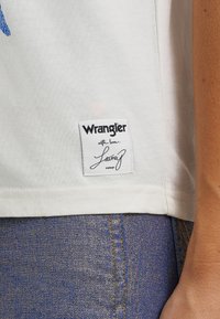 Balta kokvilnas t-krekls ar izšūtu Wrangler etiķeti, uz kuras ir rakstīts "ar mīlestību" un "Latun" kursīvā. Valkāts ar džinsiem ar zeltītām šuvēm.