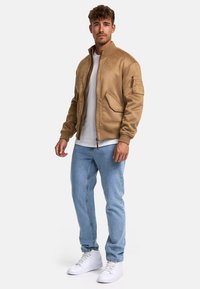 Brun bomberjakke med lynlås, ribbede ærmer og to frontlommer, parret med lyseblå jeans og hvide sneakers.