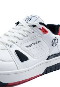 Hvit skinnsneaker med svarte og røde detaljer, med perforert design, logo på siden, og strukturert gummisåle.