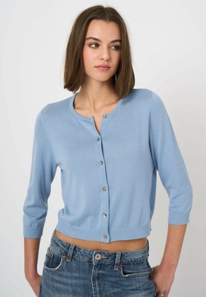 Kobieta o brązowych włosach do ramion nosi jasnoniebieski rozpinany kardigan z rękawami do łokcia oraz niebieskie jeansy z wysokim stanem.