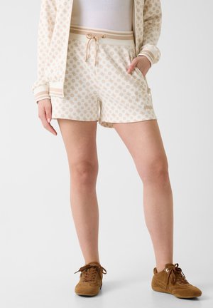 Femme portant un short beige à motifs avec cordon de serrage, une veste assortie et des chaussures décontractées marron à lacets, debout avec une main dans la poche.