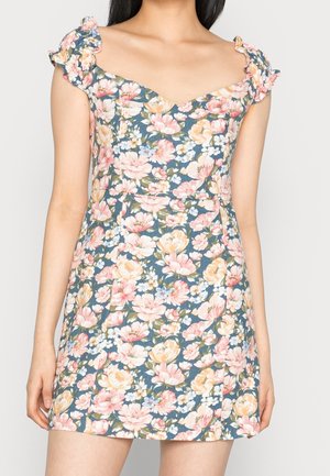 Kvinde iført en ærmeløs, kort, blomsterprintet kjole med flæsebesatte stropper og hjerteformet udskæring i pastelrosa, gul og grøn på blå baggrund.