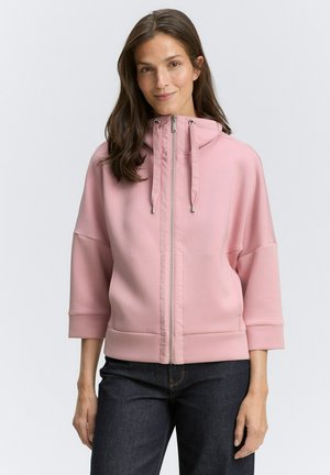 Light pink zip-up hoodie with a high collar, drawstrings, and three-quarter sleeves. Soft fabric and side pockets. Worn with dark jeans.