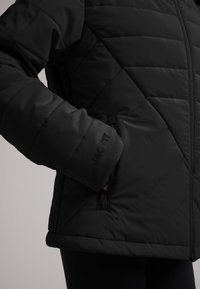 Giacca nera imbottita con un motivo a rombi, dotata di tasca con zip e logo in rilievo sul polsino. Materiale morbido e resistente all'acqua.