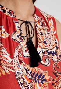 Top con motivo floreale rosso, caratterizzato da un cordoncino nero con un pon pon, scollatura dettagliata e colori vivaci in un tessuto morbido.