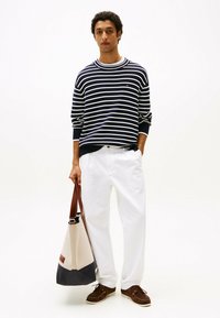 Maglione a righe blu e bianco, pantaloni bianchi, scarpe marroni con lacci e una borsa beige con un accento blu. Ensemble semplice e casual.