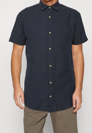 Mann trägt ein kurzärmliges, dunkelblaues Hemd mit einem dezenten Punktemuster und Khakihosen, steht vor einem schlichten Hintergrund.